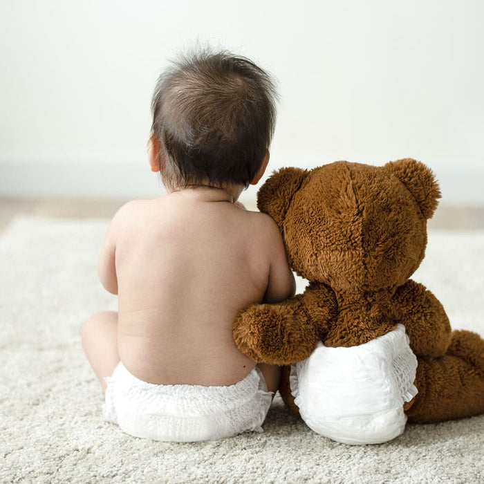 Baby sitzt mit Teddybär nebeneinander auf einem weißen Teppich